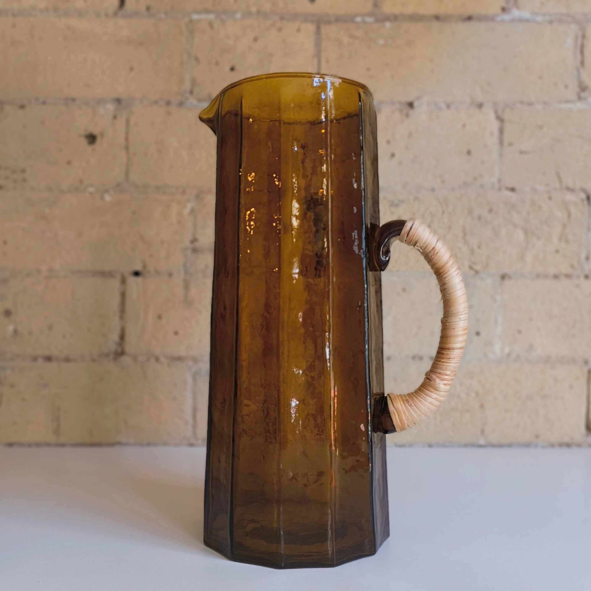 Brown glass pitcher with a handle against a brick wall.