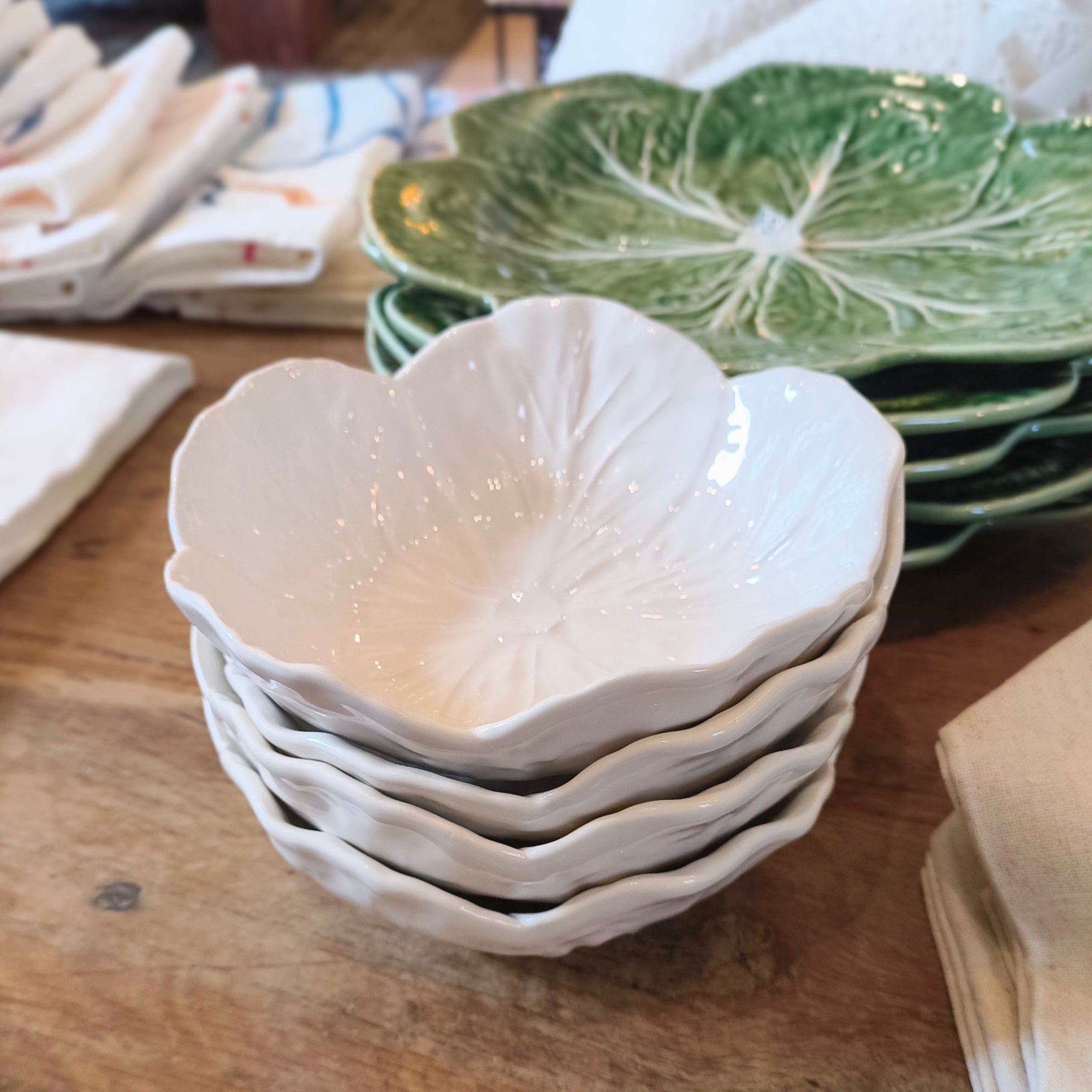 Stack of white ceramic bowls on a wooden table with green leaf-patterned plates in the background.