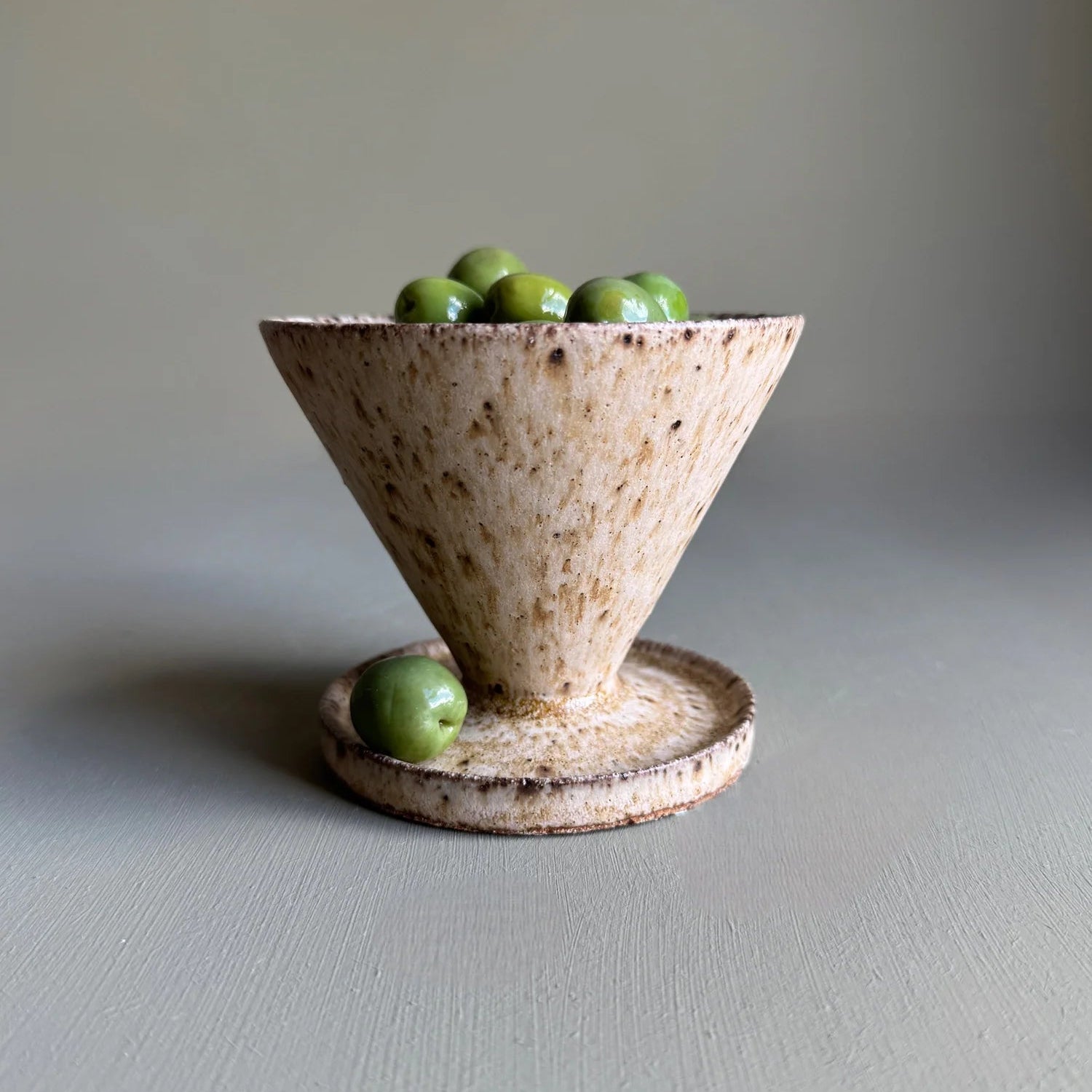 Ceramic bowl with a funnel shape containing green olives on a gray surface.