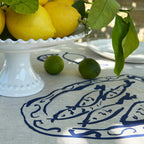 Lemons with green leaves on a white pedestal dish on a tablecloth with a fish pattern.