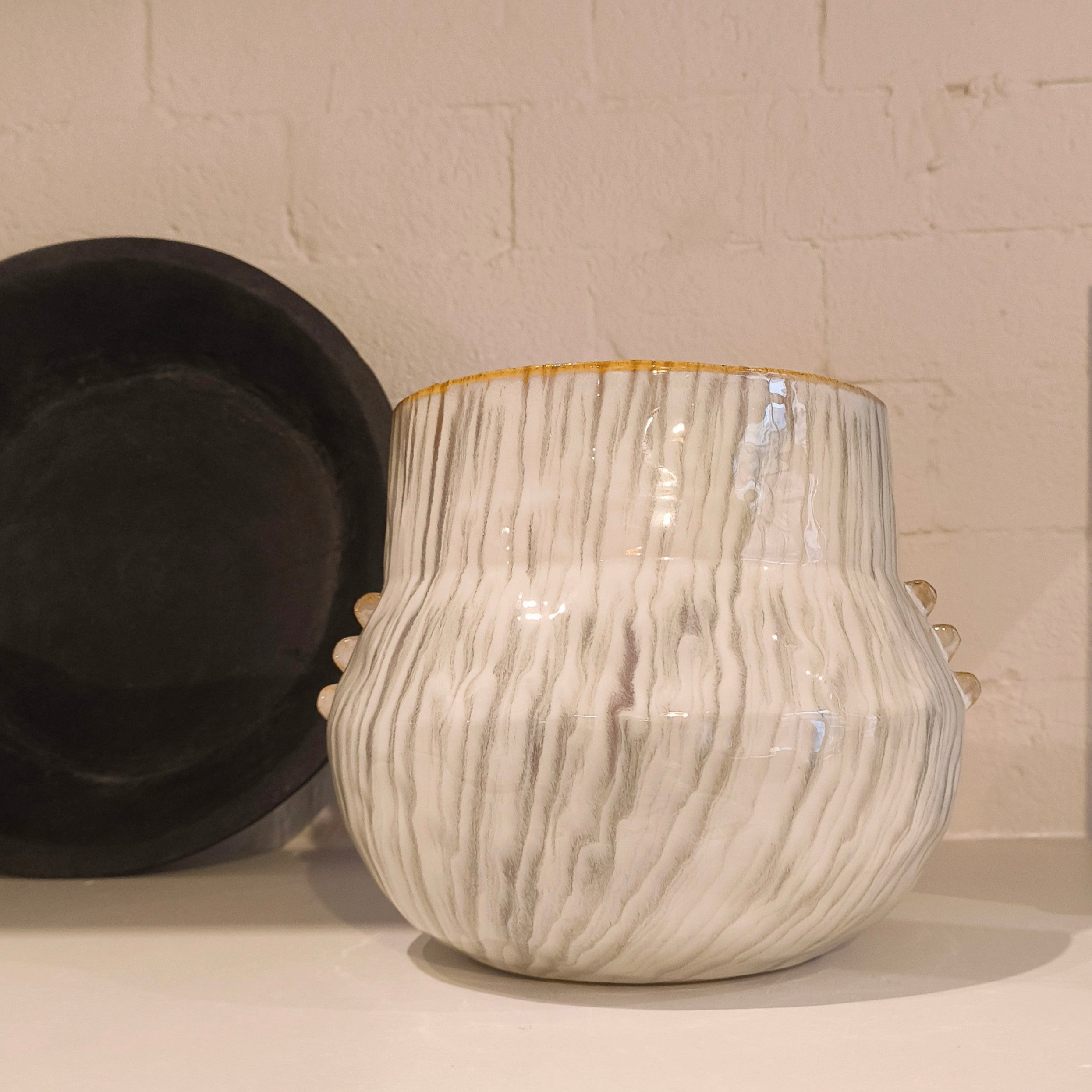 White textured vase with gold rim on a shelf against a tiled wall.