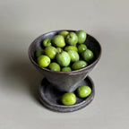 Small dark ceramic bowl filled with green olives on a light gray background