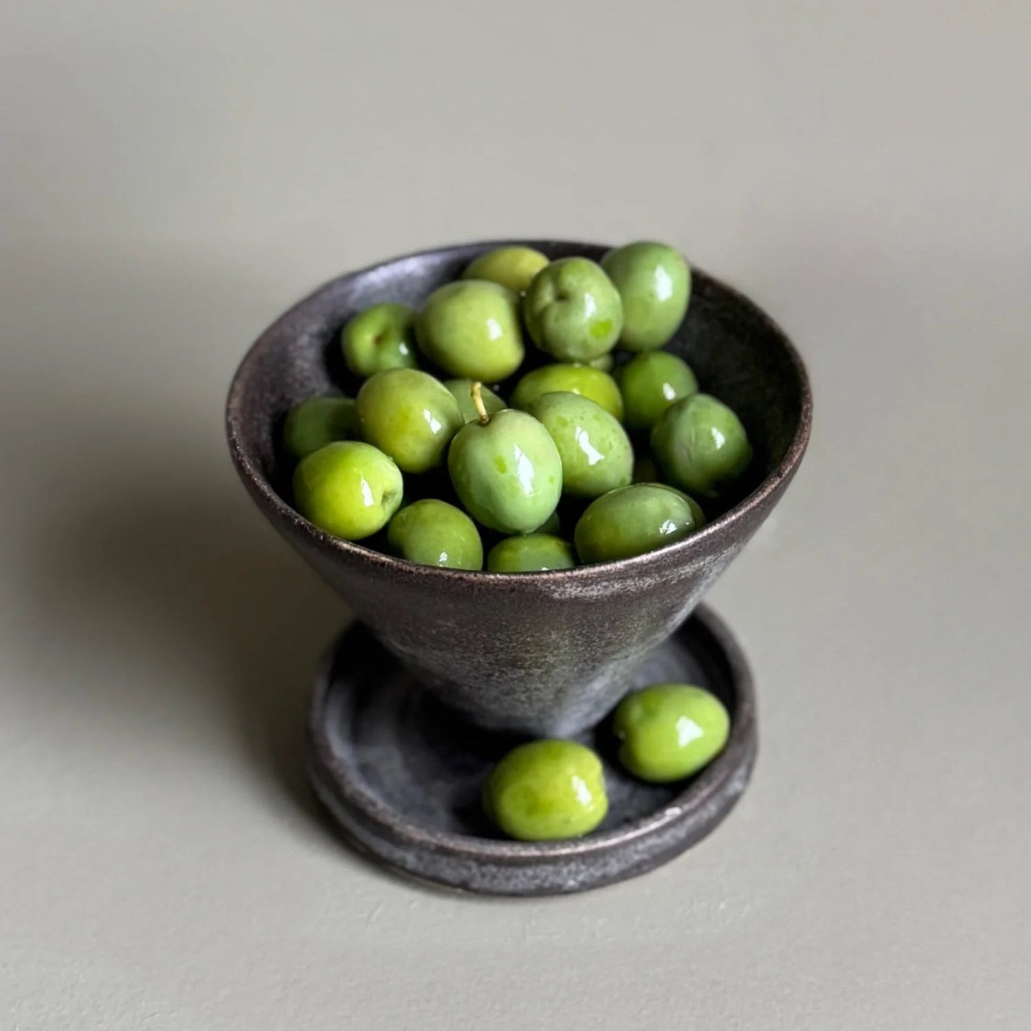 Small dark ceramic bowl filled with green olives on a light gray background