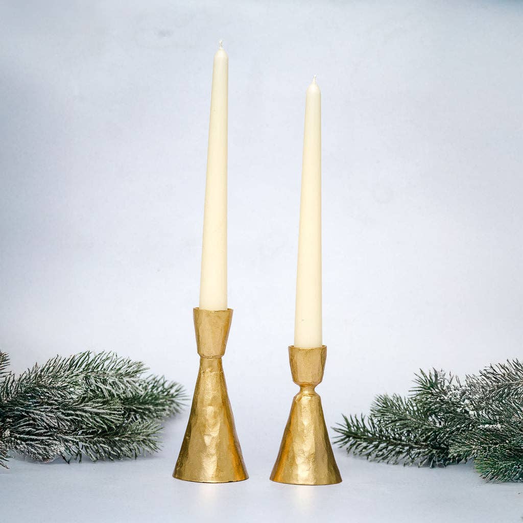 Two gold candle holders with white candles on a light gray background with decorative greenery.
