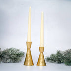 Two gold candle holders with white candles on a light gray background with decorative greenery.