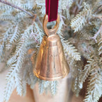 Gold bell ornament on a red ribbon hanging from a branch with white foliage.