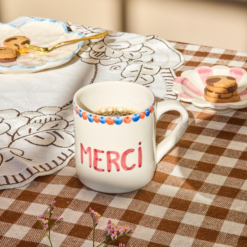Mug with 'MERCI' on a checkered tablecloth with plates and cookies in the background