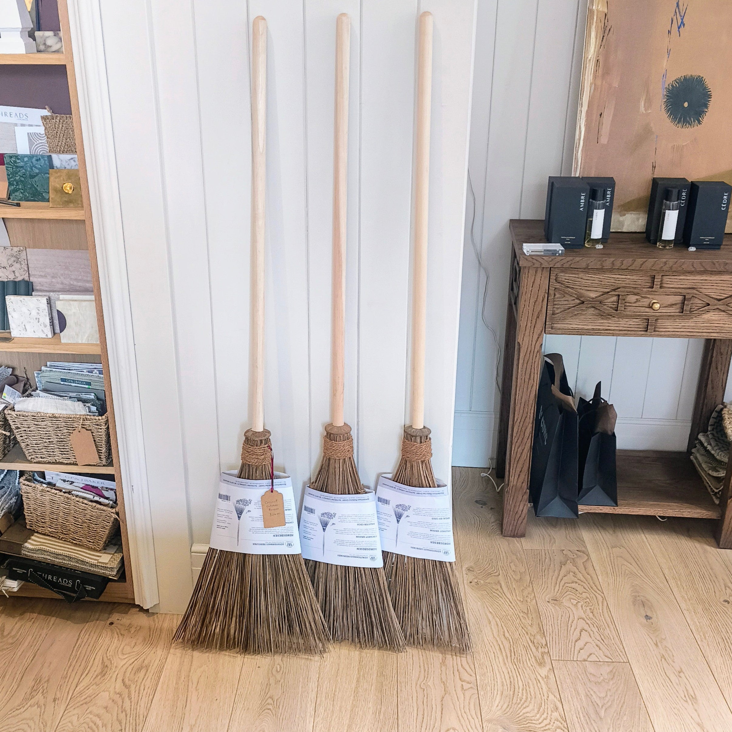 Three brooms with labels leaning against a white wall in a room with wooden flooring.