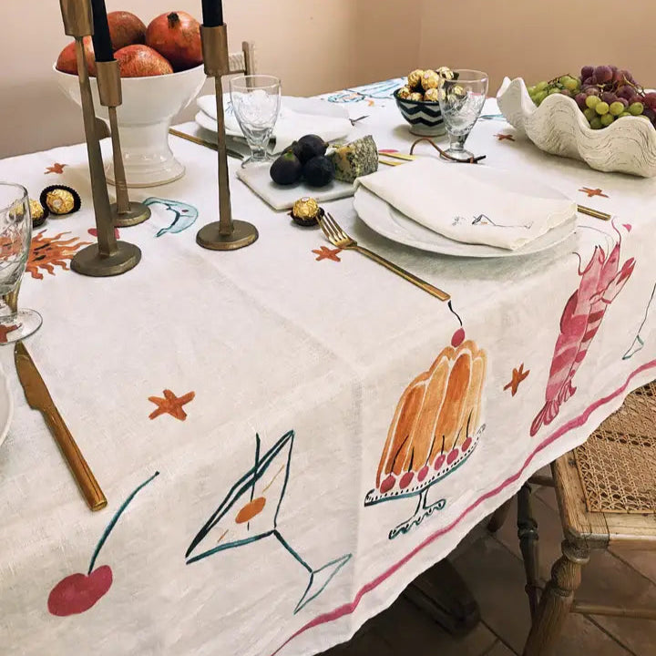 Dining table set with a decorative tablecloth featuring food illustrations, candles, and fruit.