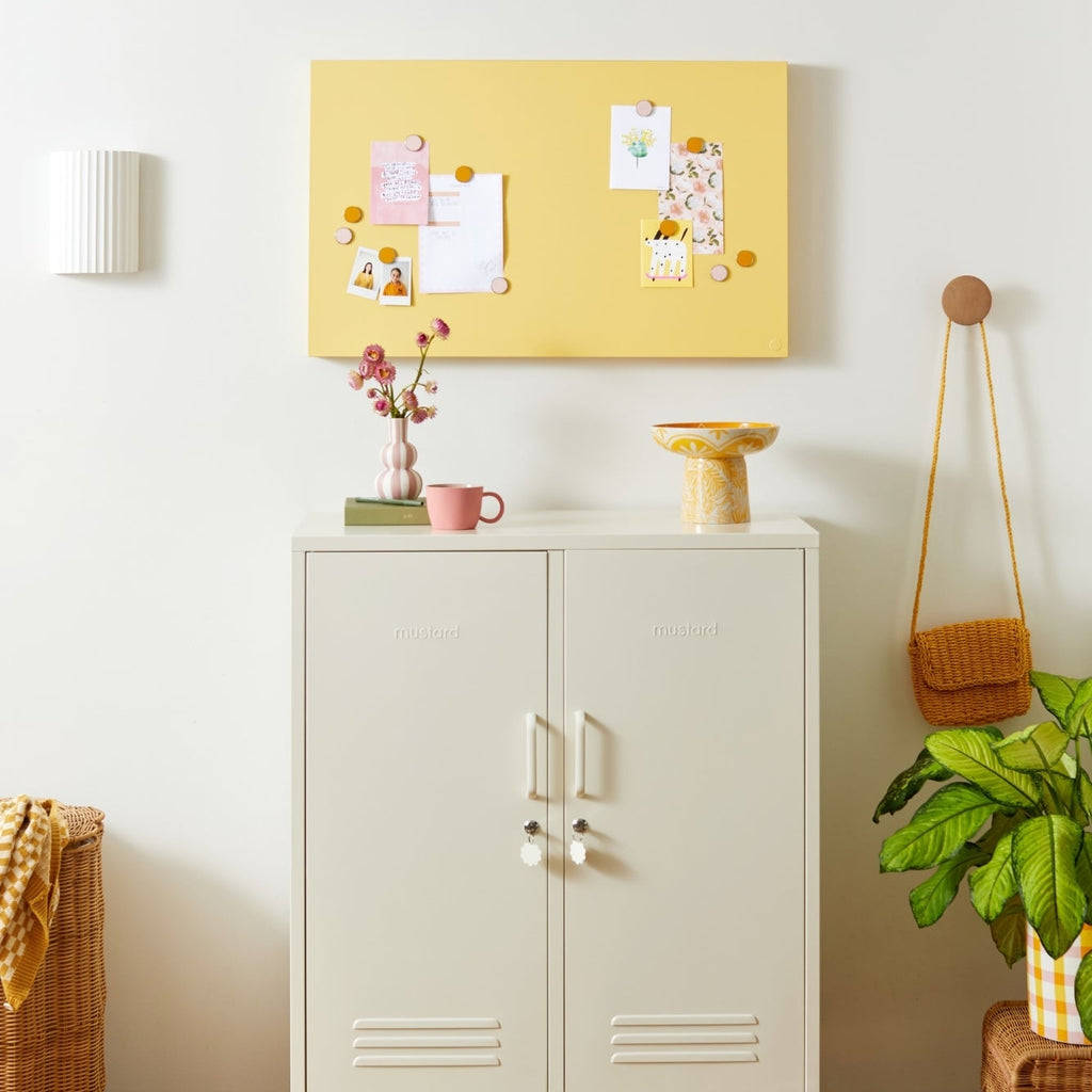 White cabinet with a yellow bulletin board above it in a room with a plant and basket.