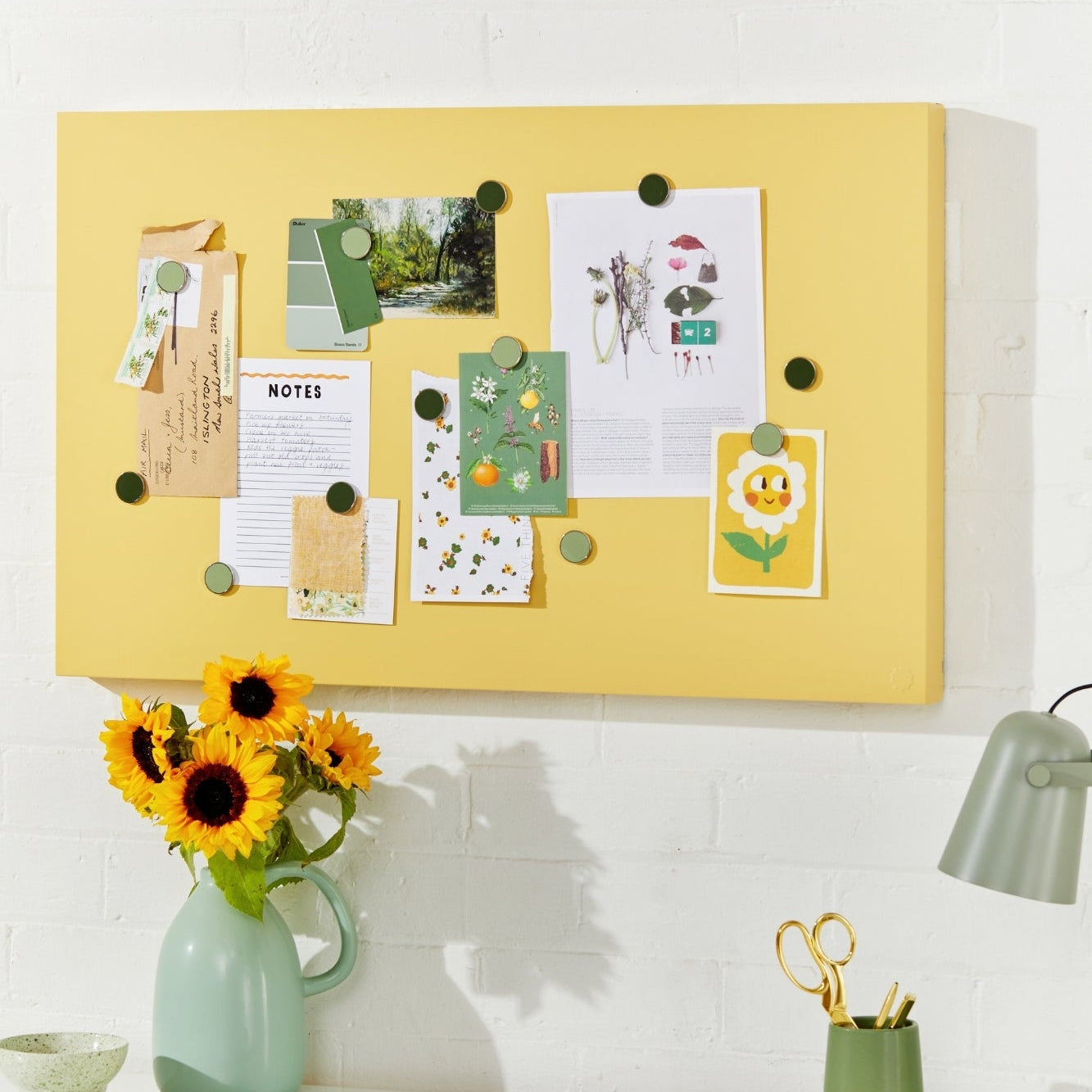 Yellow bulletin board with pinned notes above a desk with a vase of sunflowers and office supplies.