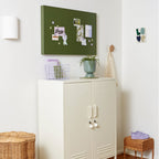 White cabinet with a green bulletin board above it in a room with wooden flooring.