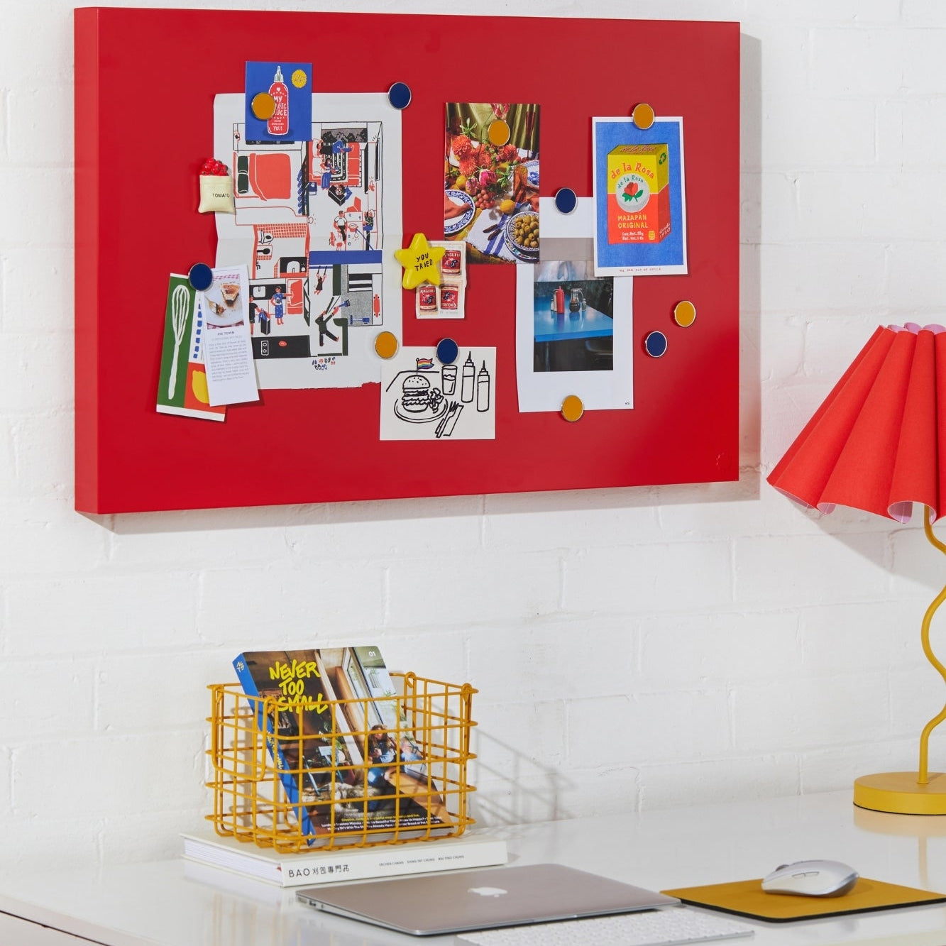 Red bulletin board with various photos and notes on a white wall above a desk.