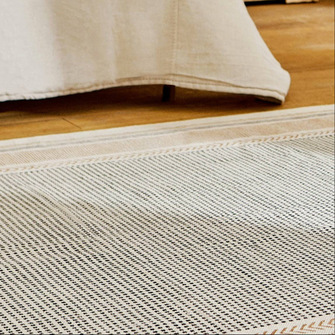 Close-up view of a flatweave rug with a hand-knotted texture, predominantly in a cream color with a rust-colored area, placed on a wooden floor beside a white curtain.