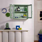 Green wall-mounted cabinet with decorative items in a room with white walls and wooden floor.