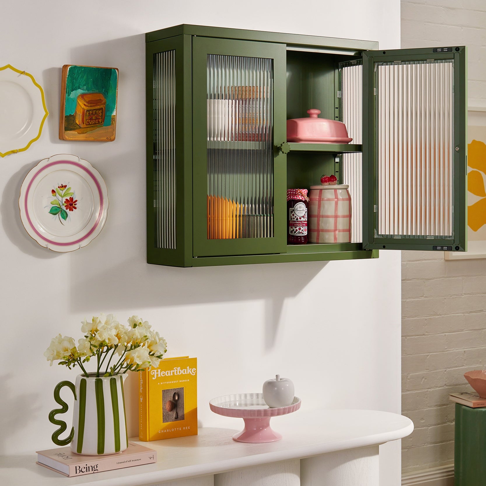 Green wall-mounted cabinet with open doors displaying items, placed on a white shelf against a white wall.