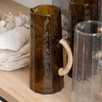 Decorative glass pitcher with handles on a kitchen counter, with a painting in the background.
