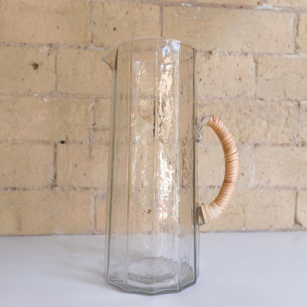 Clear glass pitcher with a wooden handle against a brick wall.