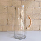 Clear glass pitcher with a wooden handle against a brick wall.