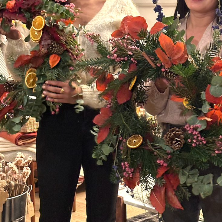 Two people holding large floral wreaths with decorative elements indoors.