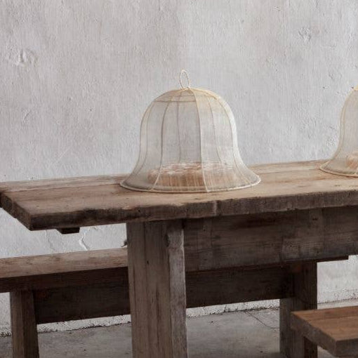 Long wooden dining table with benches with a food cover against a gray wall with hanging lamps.