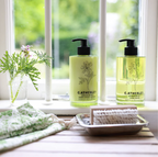 Two bottles of C. Atherley hand and body products on a windowsill with a plant and soap dish.