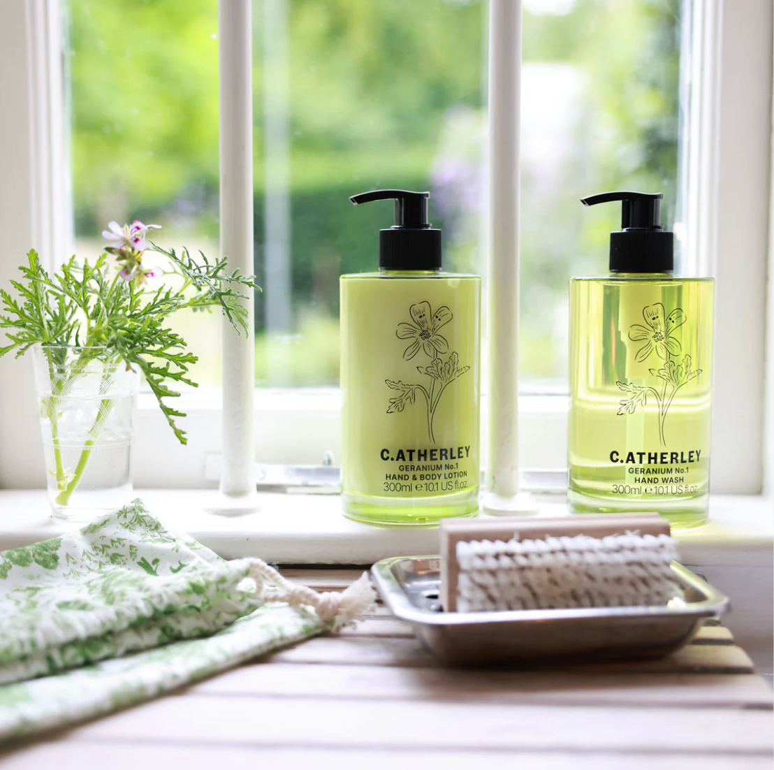 Two bottles of C. Atherley hand and body products on a windowsill with a plant and soap dish.