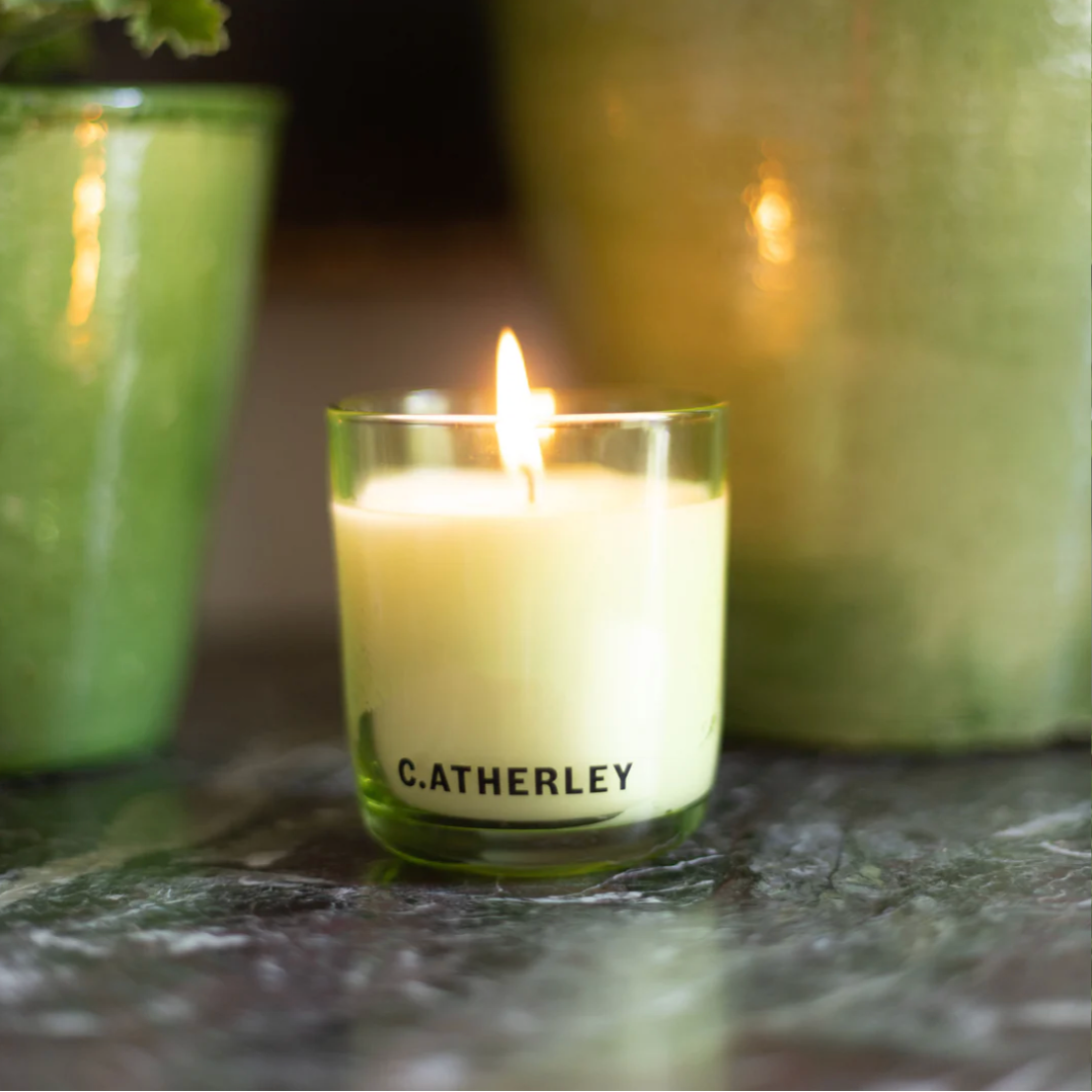 Catherley candle burning on a stone surface with green containers in the background