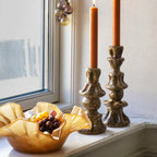 Decorative candles in ornate holders on a windowsill with a bowl of fruit.