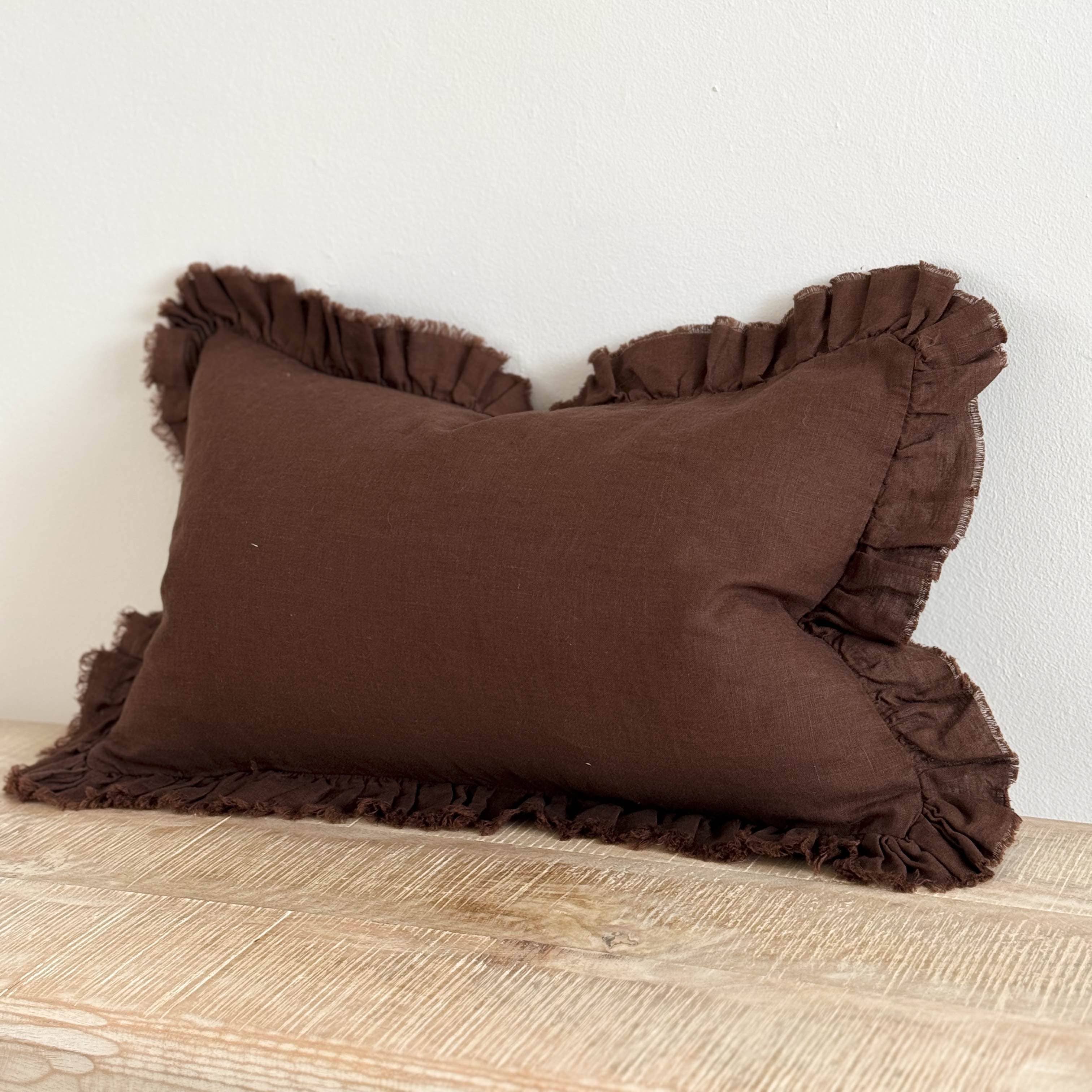 A dark brown cushion cover with a ruffled edge, displayed on a wooden surface against a white background.