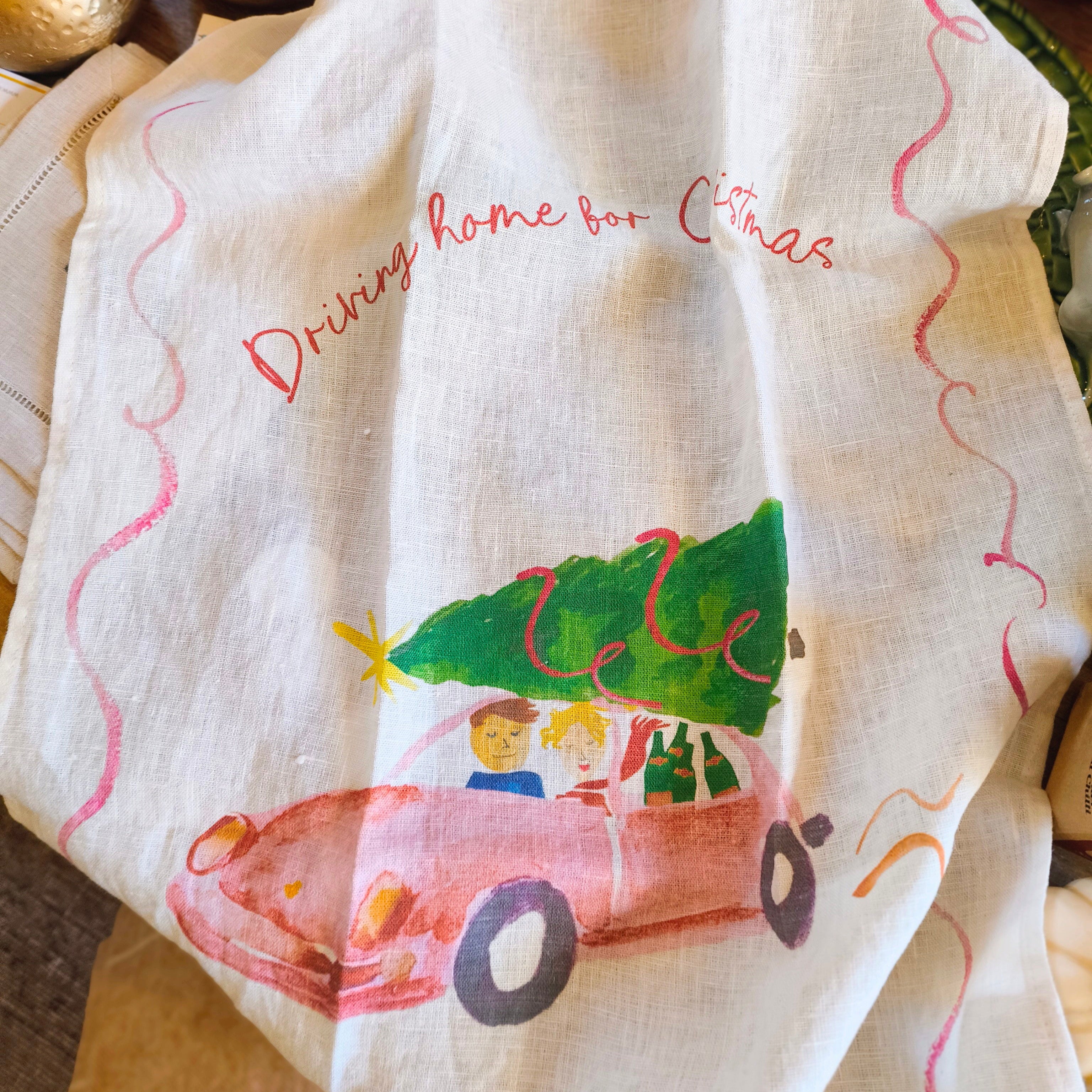 Decorative towel with a Christmas-themed design of a car carrying a tree, on a wooden surface.