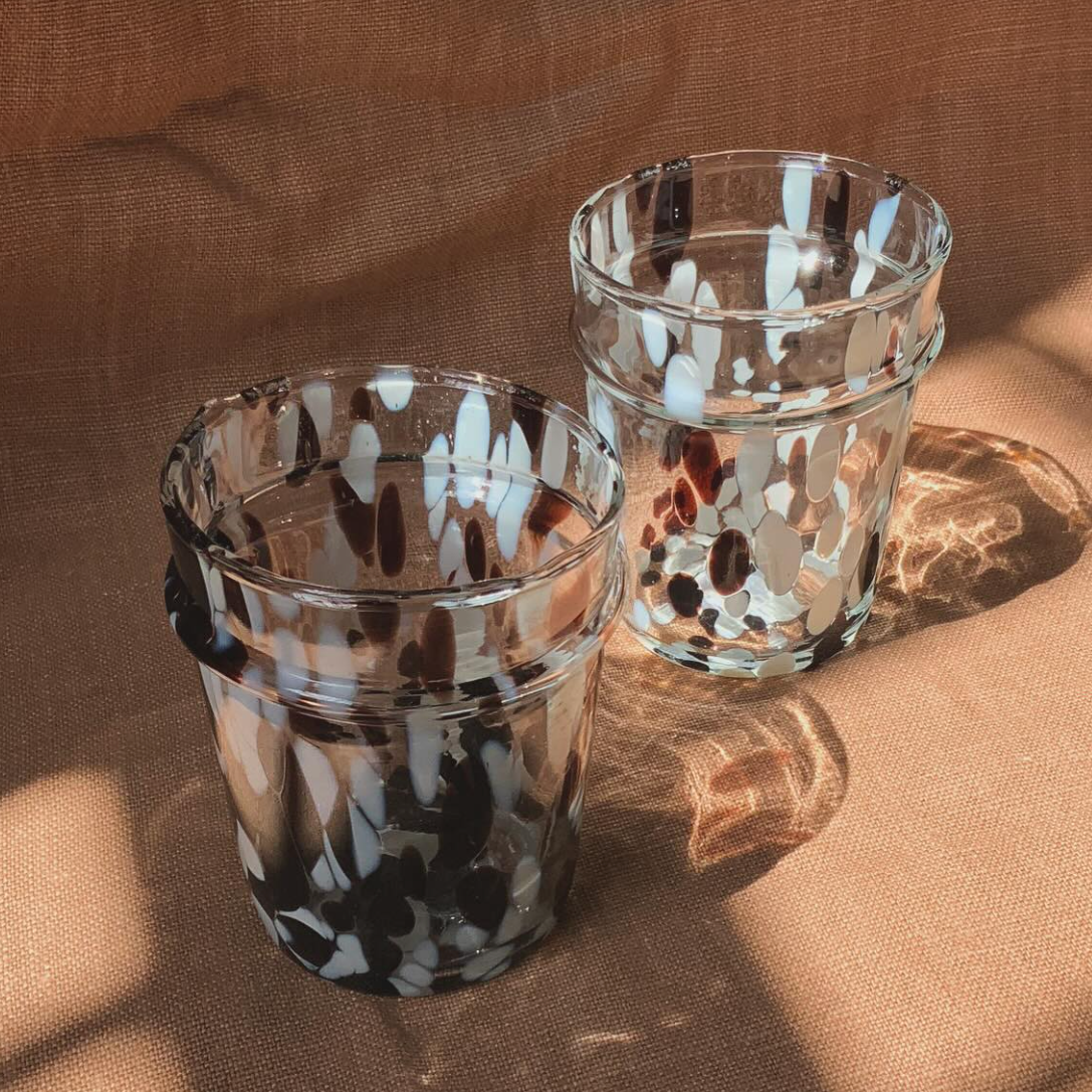 Two clear glass water glasses with a geometric pattern of brown and white spots, placed on a beige textile surface.