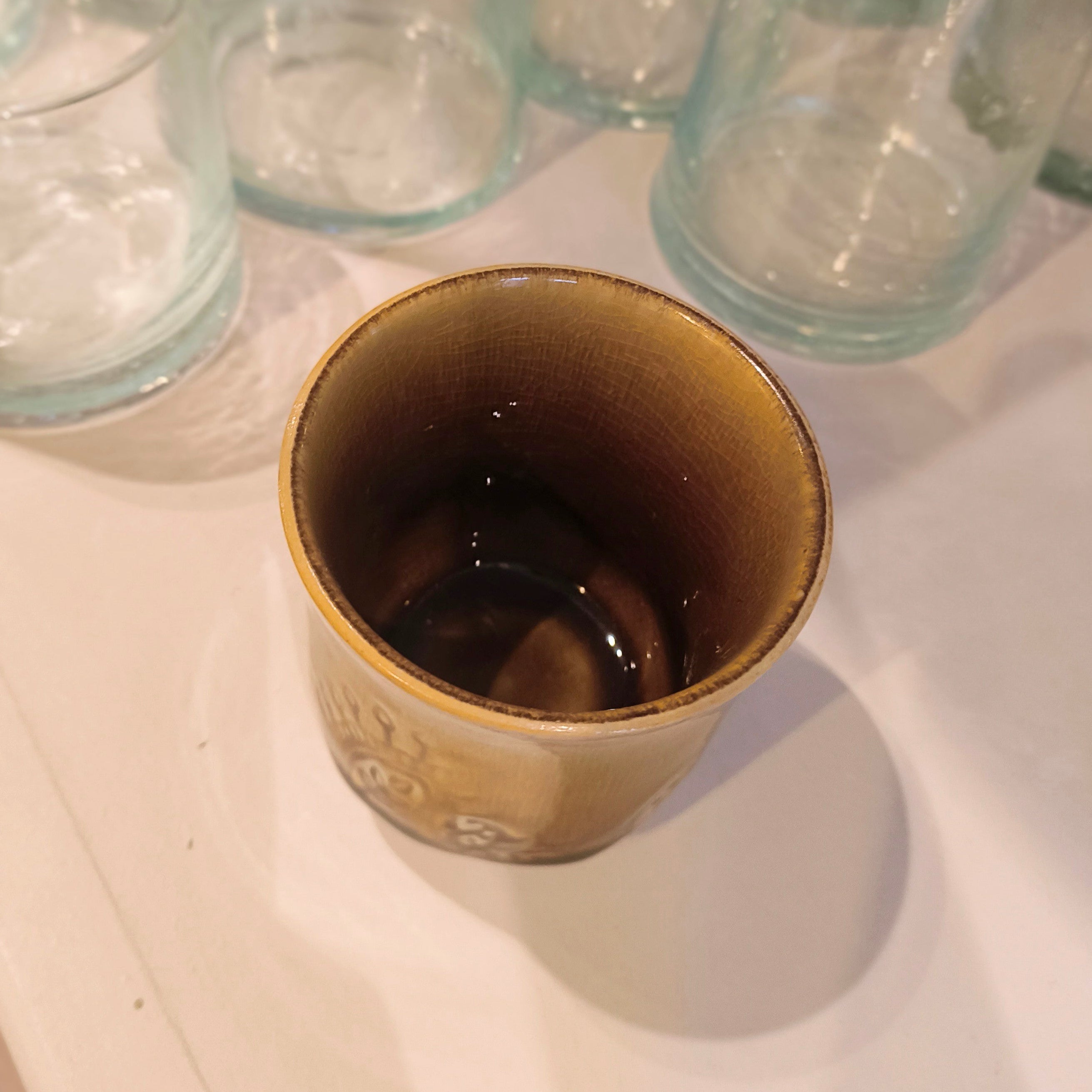 Brown ceramic cup with dark liquid on a white surface, surrounded by clear glass cups.