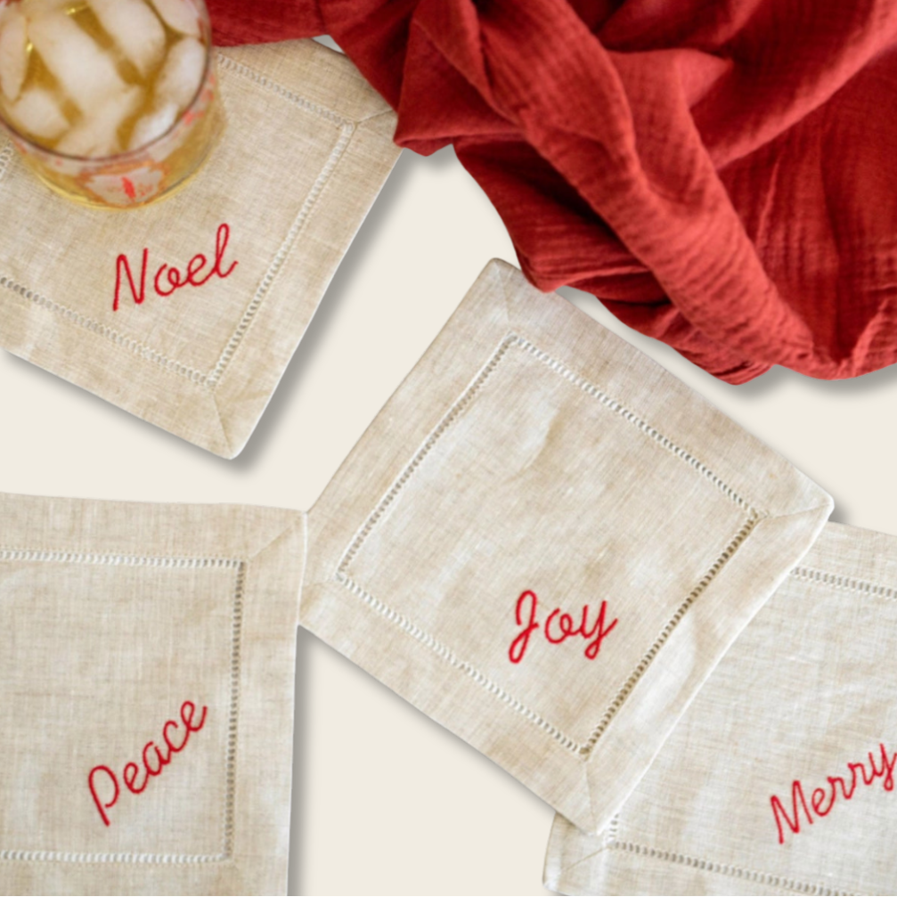 Beige napkins with red text 'Noel', 'Joy', and 'Merry' on a white surface with a red cloth and glass of iced coffee.