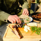 Person preparing a meal outdoors with a cutting board, pan, and ingredients.