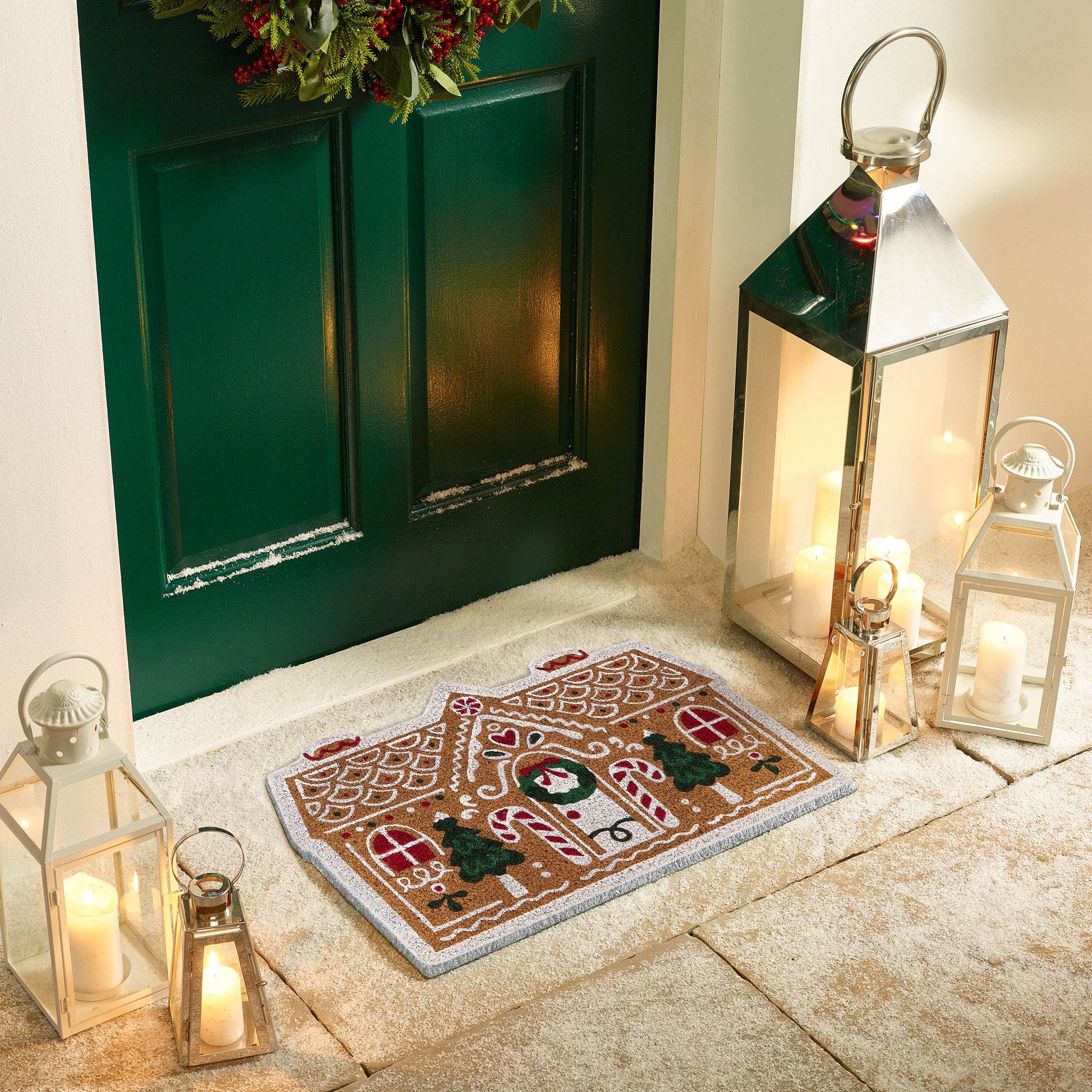 Decorative doormat with gingerbread house design in front of a green door with lanterns and candles.
