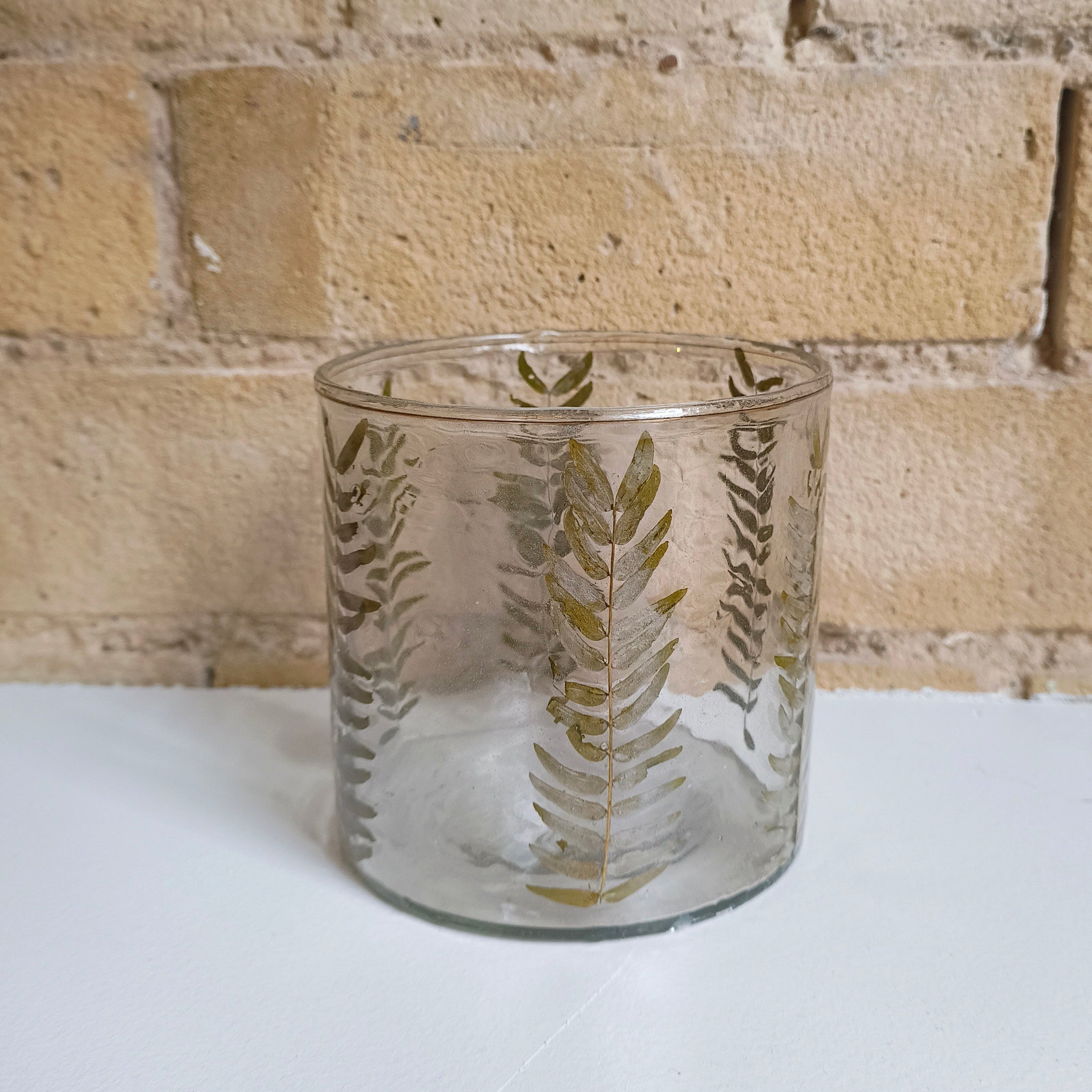 Clear glass candle holder with leaf patterns on a white surface and brick wall background