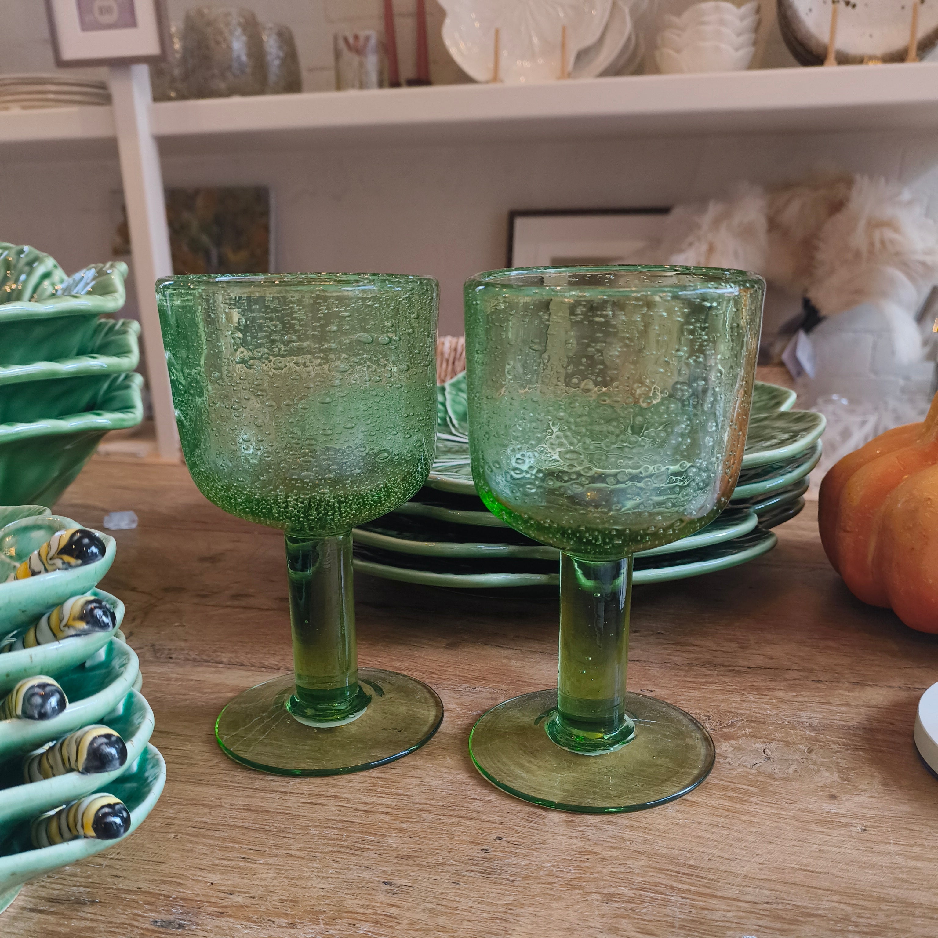 Two green glass goblets on a wooden table with decorative items in the background.