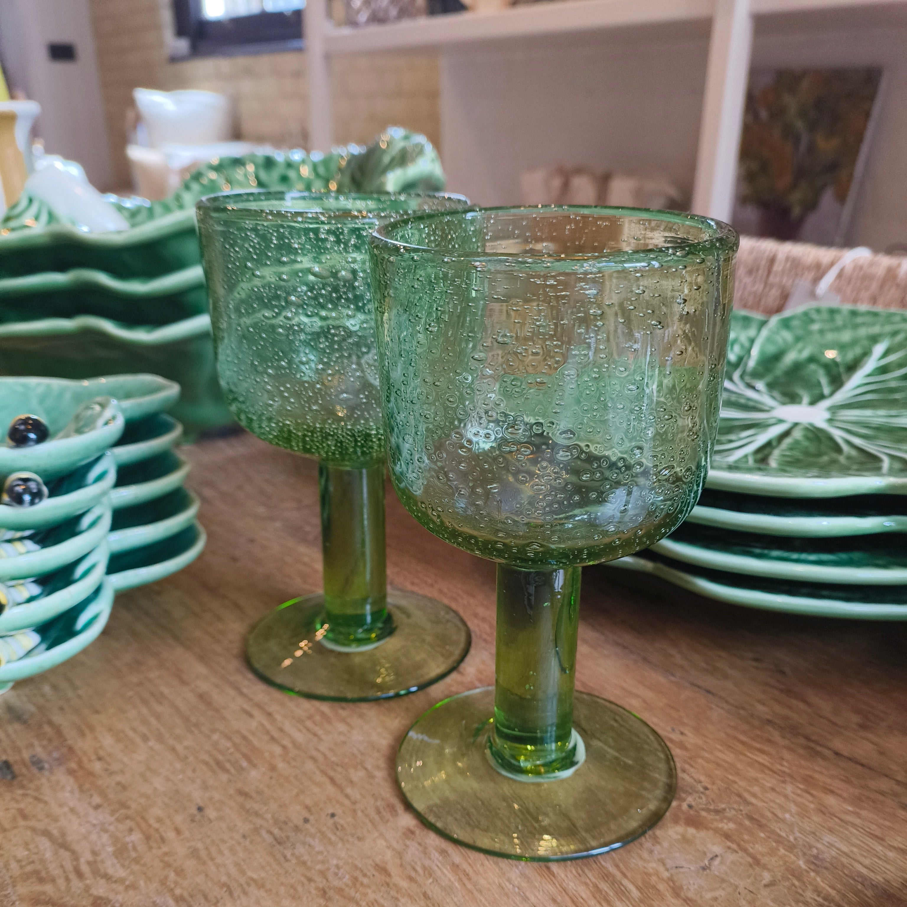 Two green glass goblets on a wooden table with plates in the background.