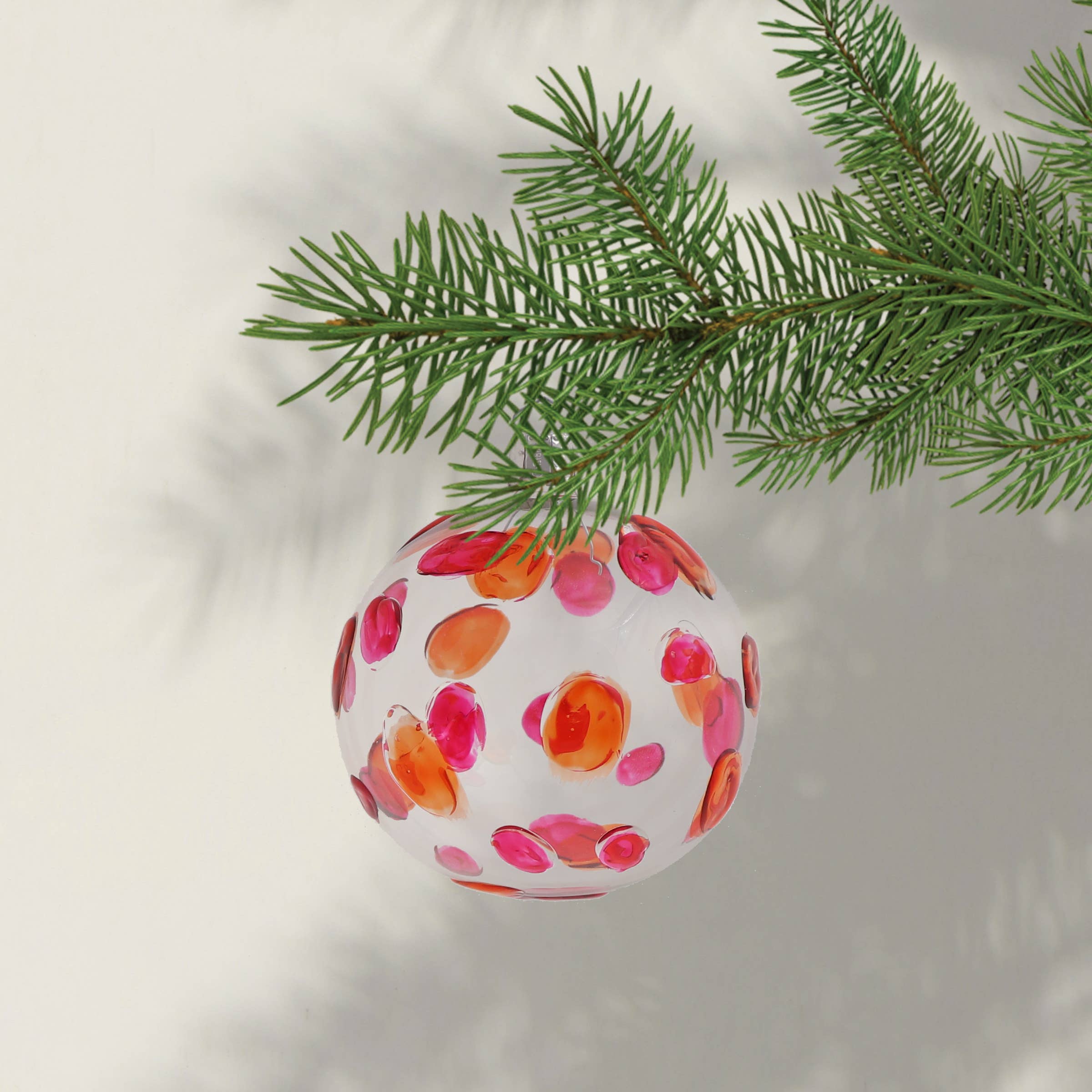 A pink and orange spotty Christmas bauble hanging from a festive tree branch.