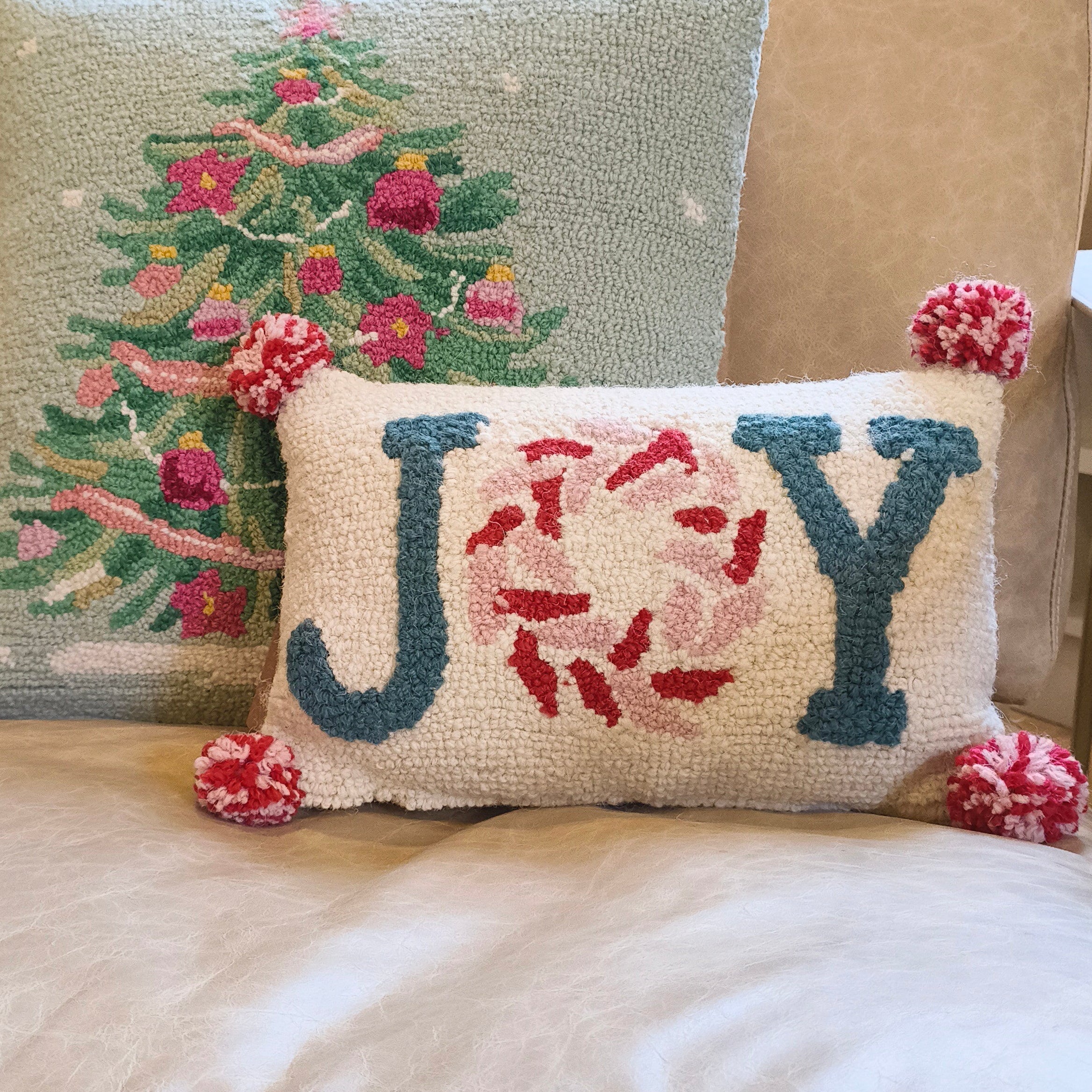 Two decorative pillows on a beige chair, one with a Christmas tree design and the other with 'JOY' letters.