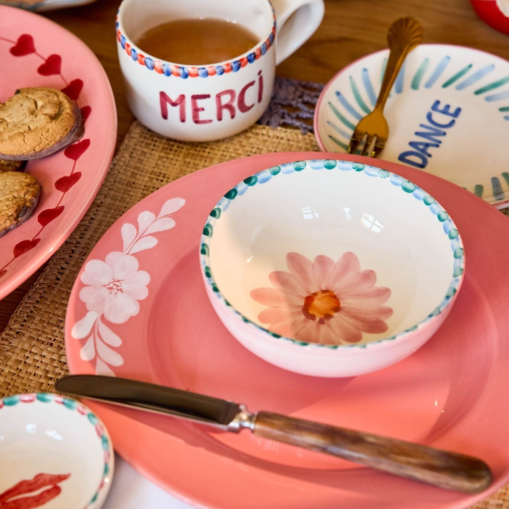 Table setting with pink plates, floral bowls, and a 'Merci' mug on a woven placemat.