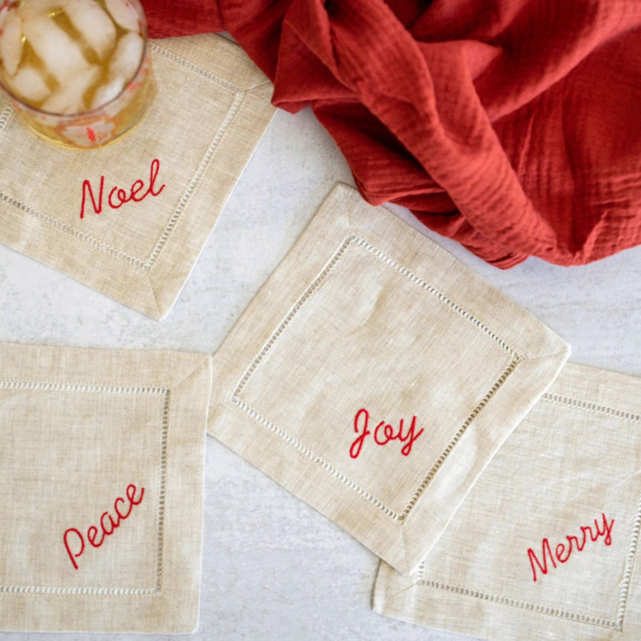 Beige napkins with red text 'Noel', 'Joy', and 'Merry' on a white surface with a red cloth and glass of iced coffee.