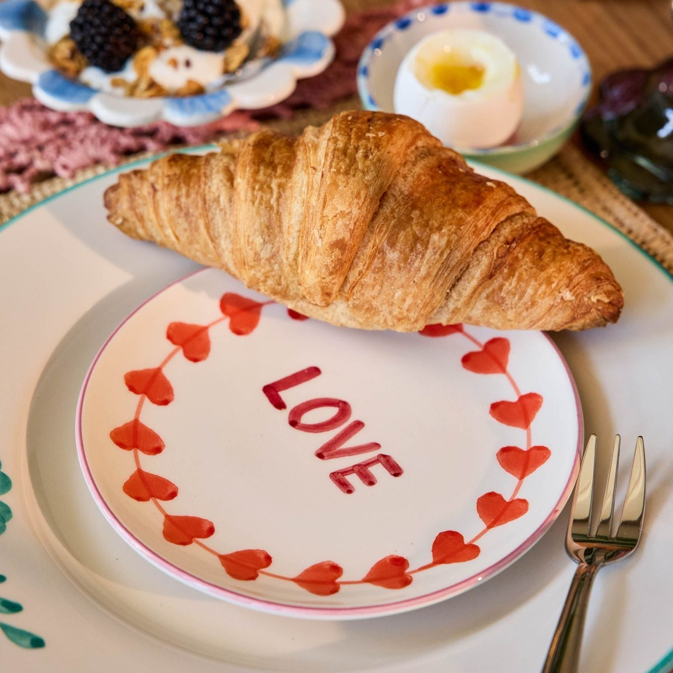 Breakfast scene with a croissant on a 'LOVE' plate, pink mug, and fruit salad.