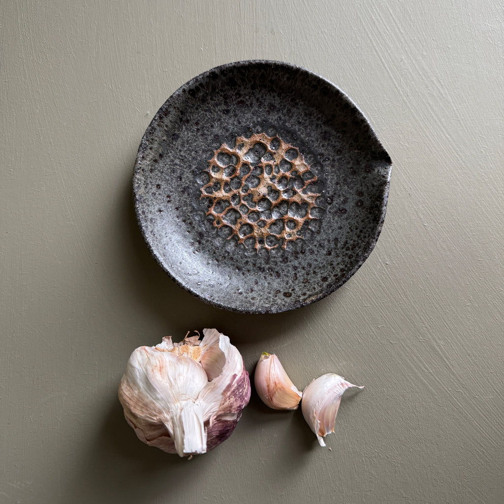Decorative black ceramic plate with a textured design on a green background, accompanied by garlic bulbs.