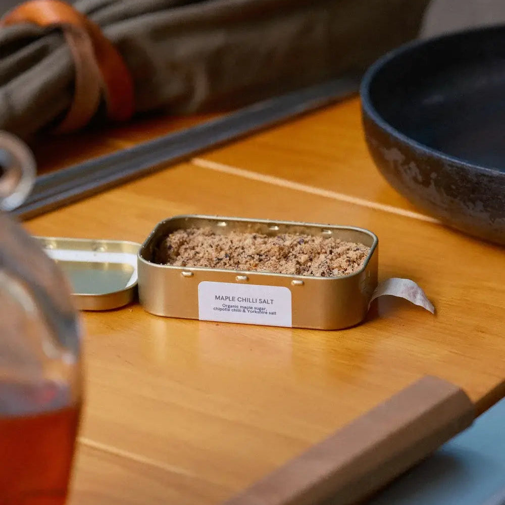 Small metal container with food on a wooden table with a glass of tea and other items.