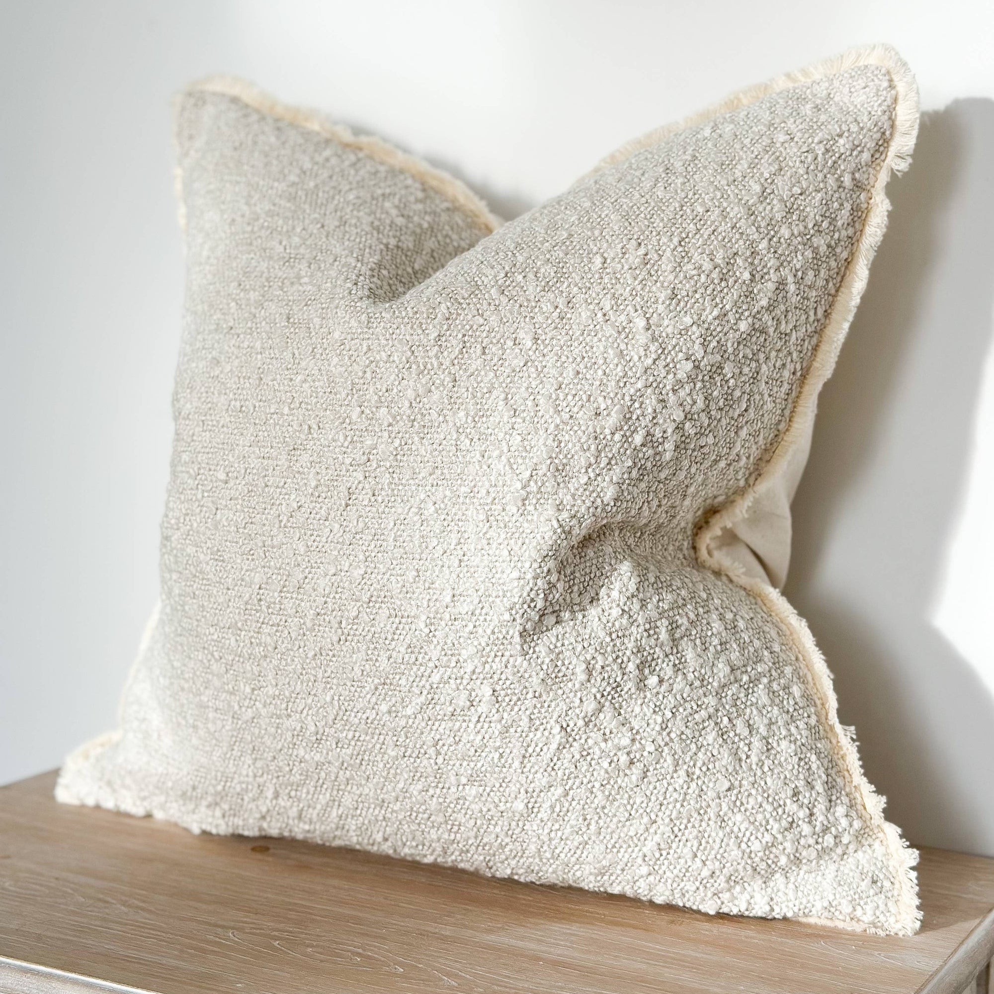 A cream-colored boucle cushion cover with a fringed edge on a wooden surface against a white wall.