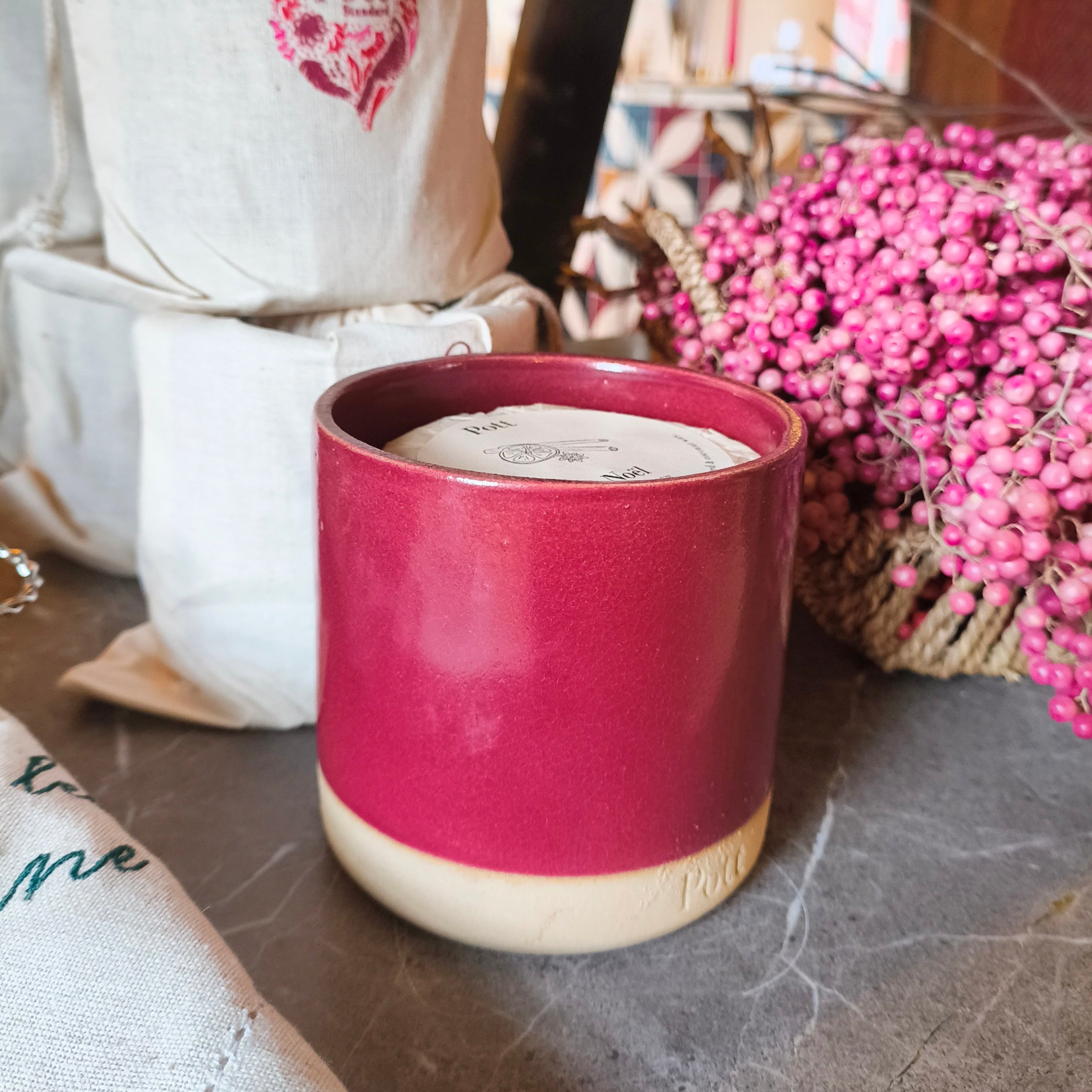 Pink ceramic container with a beige base on a marble surface, with a white bag and pink flowers in the background.