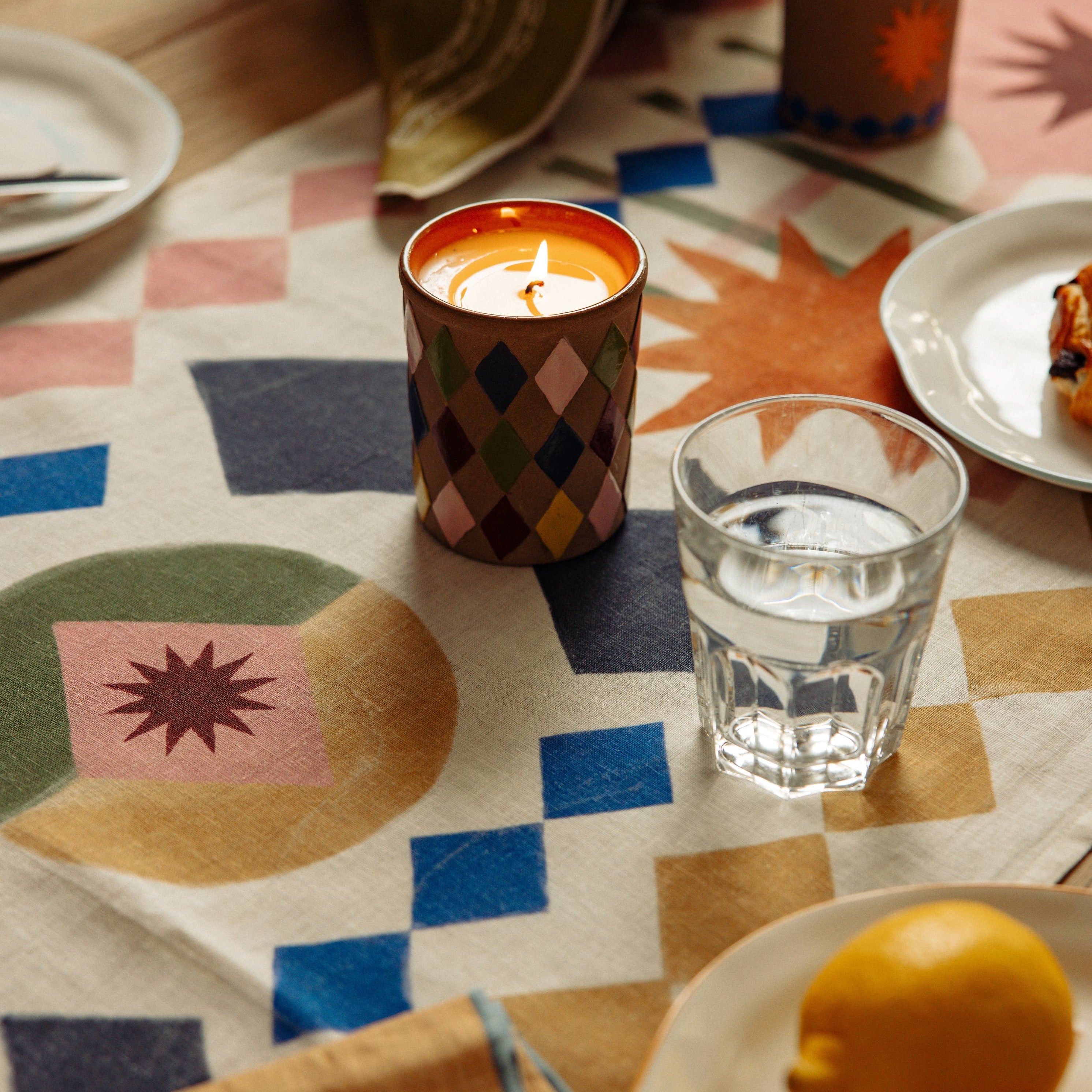 Dining table setting with candles, a glass of water, and a lemon on a patterned tablecloth.