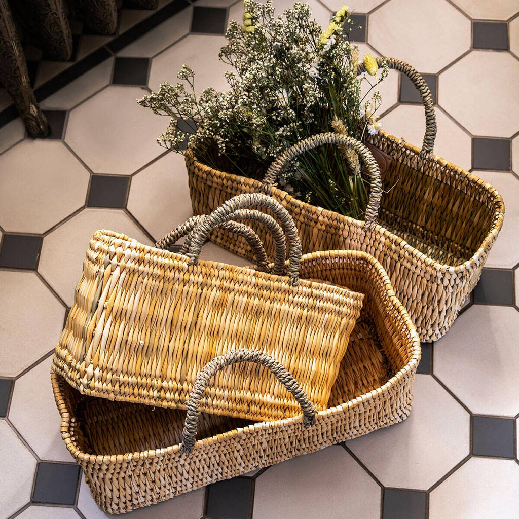 Traditional Moroccan reed baskets with sisal handles, placed on a tiled floor. One basket contains flowers, and the others are empty.
